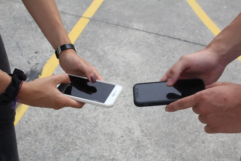 Two people holding smartphones facing each other outdoors, as if comparing the two phones side by side.