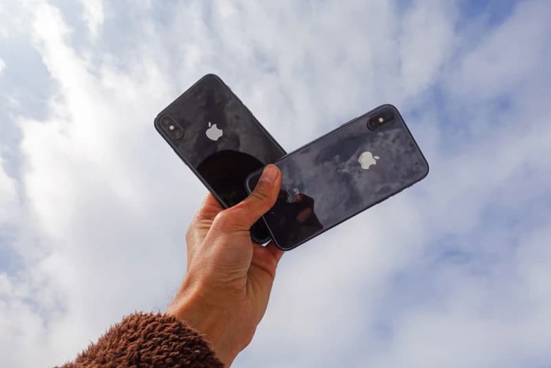 A hand holds up two black smartphones against a cloudy sky.